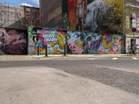 a wall with different colorful artwork on it in a city with buildings around and the street has a bike path going under it