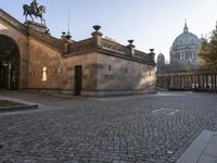 people ride horses in a courtyard near buildings, while buildings are behind them with columns