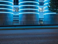 blue lights glowing up on two cylindrical buildings and a tree in front of a road