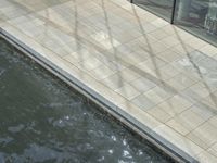 water is shown along a sidewalk with a bench next to it and buildings in the background