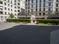 a small paved courtyard in front of some white buildings with green hedges and trees on both sides