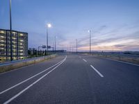 a paved highway next to a tall building at dusk time with street lights nearby the road