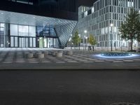 a paved area in front of tall buildings with lights on them at night with blue water spewing from the street