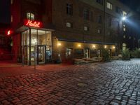 a stone sidewalk that has red neon signs on it in front of a building that has a restaurant next to it