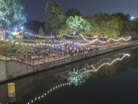 the people are enjoying an outdoor eating event near a river at night with lights strung from trees