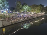 the people are enjoying an outdoor eating event near a river at night with lights strung from trees