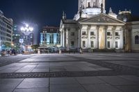 a building with a clock tower in the middle of the square at night time a man with a suitcase stands next to it