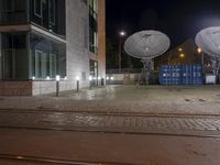 two satellite dishes suspended over a street next to a curb and building at night in the background