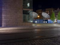 two satellite dishes suspended over a street next to a curb and building at night in the background