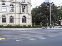 three people ride bikes past an old style building, with several tall trees and bushes