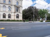 three people ride bikes past an old style building, with several tall trees and bushes