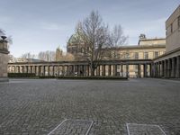 a picture of a courtyard with a few columns on the left and a small garden with benches in front