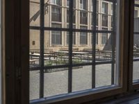 an outdoor patio with tables and benches, viewed through a window in a building with brick walls, has a bench outside