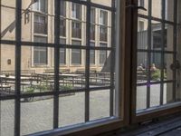 an outdoor patio with tables and benches, viewed through a window in a building with brick walls, has a bench outside