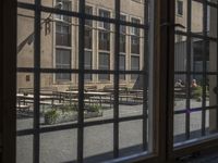 an outdoor patio with tables and benches, viewed through a window in a building with brick walls, has a bench outside