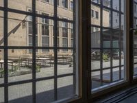 an outdoor patio with tables and benches, viewed through a window in a building with brick walls, has a bench outside