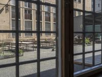 an outdoor patio with tables and benches, viewed through a window in a building with brick walls, has a bench outside