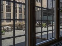 an outdoor patio with tables and benches, viewed through a window in a building with brick walls, has a bench outside