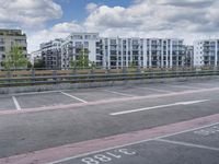 a photo of an empty parking lot surrounded by tall buildings and a fence, from which has a blue sky and some clouds