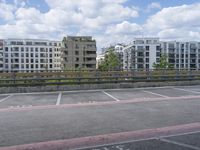 a photo of an empty parking lot surrounded by tall buildings and a fence, from which has a blue sky and some clouds