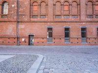 the building is made of red brick with doors and windows there is a man walking down a cobble stone sidewalk