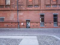 the building is made of red brick with doors and windows there is a man walking down a cobble stone sidewalk