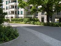 a stone street in front of a tall building with many plants around it and several bushes outside