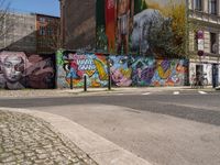 a wall with different colorful artwork on it in a city with buildings around and the street has a bike path going under it