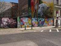 a wall with different colorful artwork on it in a city with buildings around and the street has a bike path going under it