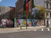 a wall with different colorful artwork on it in a city with buildings around and the street has a bike path going under it