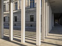 several pillars, one with shadows, are in a courtyard with windows and a tall white building behind them