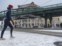 a woman is running on a snowy street in front of a bridge over a crowded city area