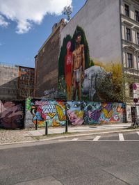 a wall with different colorful artwork on it in a city with buildings around and the street has a bike path going under it