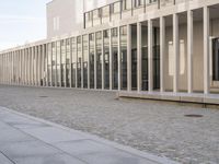 a brick pathway leading to a white building with large windows and several columns on the side