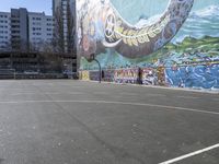 a basketball court with graffiti on the side and a tree beside it and in the distance, an apartment building behind