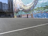 a basketball court with graffiti on the side and a tree beside it and in the distance, an apartment building behind