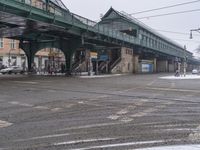 a green pedestrian bridge and cars passing under it on a snowy day, with a sidewalk in the foreground