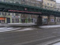 a street with snow in the foreground under a bridge and buildings in the background