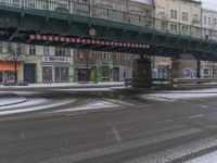 a street with snow in the foreground under a bridge and buildings in the background