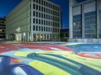 colorful street art on a street in front of an office building at night - shaped like an arch, with a water fountain