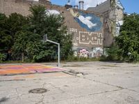 an abandoned street with a mural on it's side, near trees and a basketball court