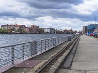 the sidewalk near the water is being made of stone, and there are train tracks leading to a dock where cars sit parked