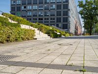 a brick city sidewalk with a stone curb, next to a building, in a park or courtyard, in front of a park, surrounded by bushes and some stairs