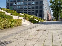 a brick city sidewalk with a stone curb, next to a building, in a park or courtyard, in front of a park, surrounded by bushes and some stairs