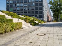 a brick city sidewalk with a stone curb, next to a building, in a park or courtyard, in front of a park, surrounded by bushes and some stairs