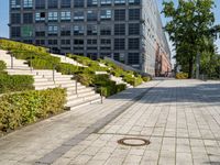 a brick city sidewalk with a stone curb, next to a building, in a park or courtyard, in front of a park, surrounded by bushes and some stairs