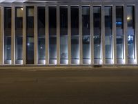 a man on a sidewalk standing in front of a building with some windows open at night