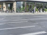 two people walking across the road past a building that has several bikes in it and two parked bikes on the sidewalk