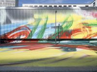 a woman walking past a mural painted on a fence in a city street area on a bench with a bright colored blanket in the sunlight