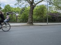 two people riding bikes on a road in an open city area next to trees and bushes